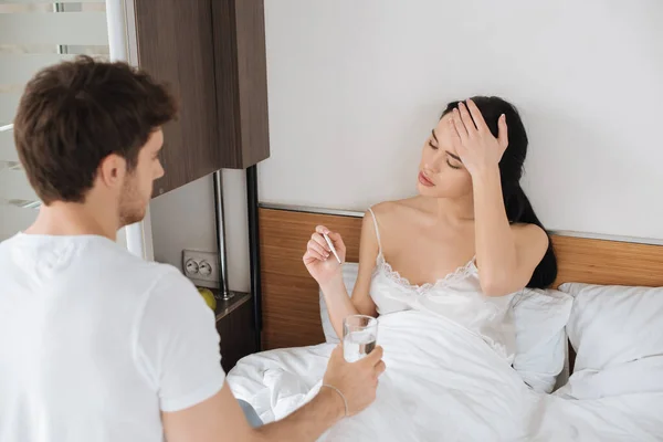 Man giving glass of water to ill girlfriend with thermometer in bed during self isolation — Stock Photo