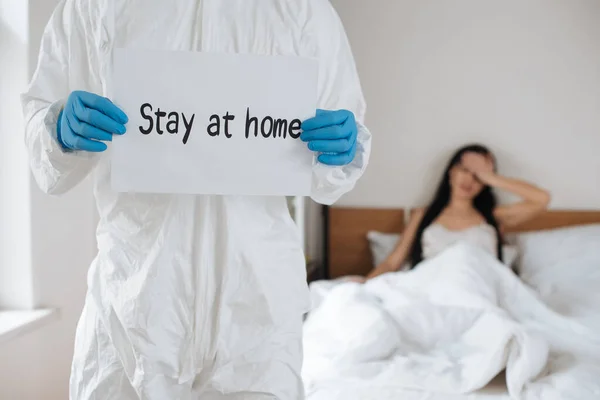 Cropped view of doctor in protective suit holding card with Stay at home lettering in bedroom with ill woman, selective focus — Stock Photo
