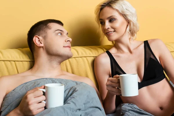 Smiling couple looking at each other in bed under blanket with mugs on yellow background — Stock Photo