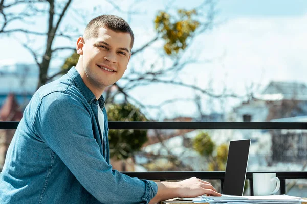 Smiling freelancer sitting on terrace with laptop and papers — Stock Photo