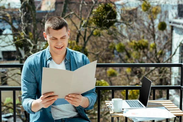 Freelancer sorridente sentado no terraço com pasta perto de laptop e documentos — Fotografia de Stock