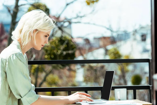 Vista lateral del freelancer rubio sentado en la terraza y usando el ordenador portátil - foto de stock