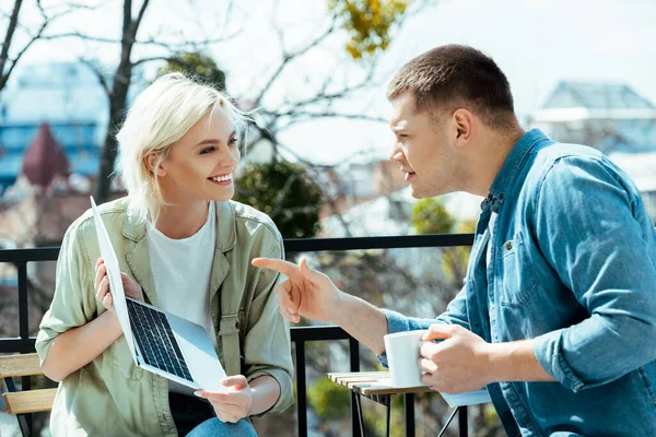 Mann mit Becher zeigt auf Laptop-Bildschirm neben lächelnder Frau auf sonniger Terrasse — Stockfoto