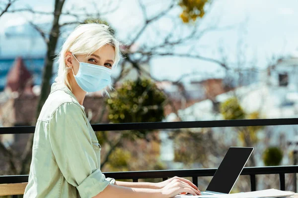 Вид збоку блондинки фрілансер в медичній масці, що сидить на терасі і використовує ноутбук — стокове фото