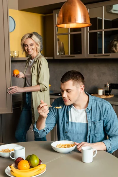 Donna sorridente in piedi vicino al marito mangiare cornflakes in cucina — Foto stock