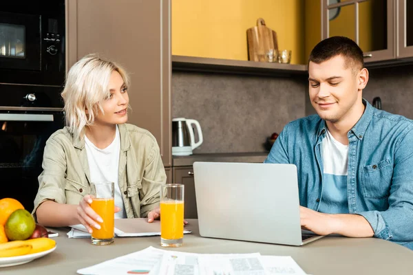 Sonriente pareja trabajando con papeles y portátil cerca de jugo de naranja en gases en la cocina en casa - foto de stock