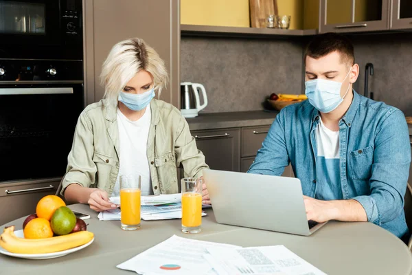 Couple dans des masques médicaux travaillant avec des papiers et un ordinateur portable près de jus d'orange dans les gaz dans la cuisine à la maison — Photo de stock