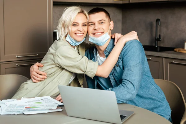 Щаслива пара в медичних масках обіймається під час роботи з паперами та ноутбуком на кухні вдома — стокове фото