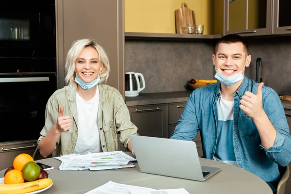 Happy couple showing thumbs up in medical masks working with papers and laptop in kitchen at home — Stock Photo