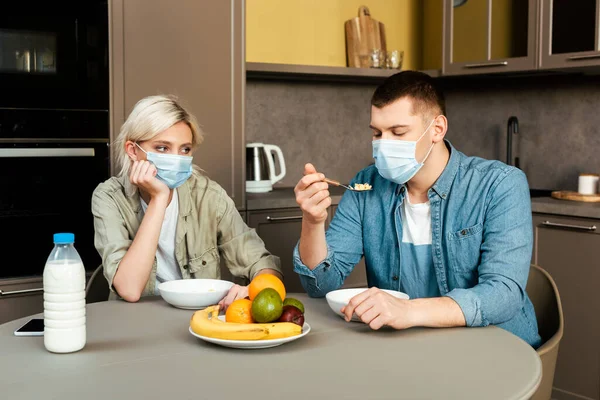 Pareja en máscaras médicas desayunando en la cocina - foto de stock