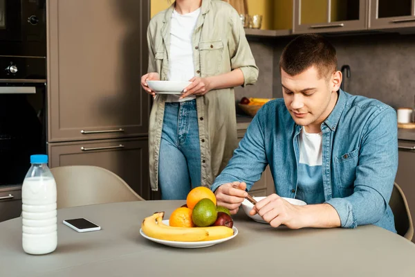 Vista cortada da mulher de pé perto do homem tomando café da manhã à mesa com leite e frutas na cozinha — Fotografia de Stock