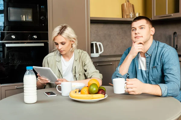 Женщина, использующая цифровой планшет рядом мечтательный муж во время завтрака на кухне — стоковое фото