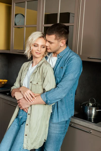 Sonriente joven pareja amorosa abrazándose en la cocina en casa - foto de stock