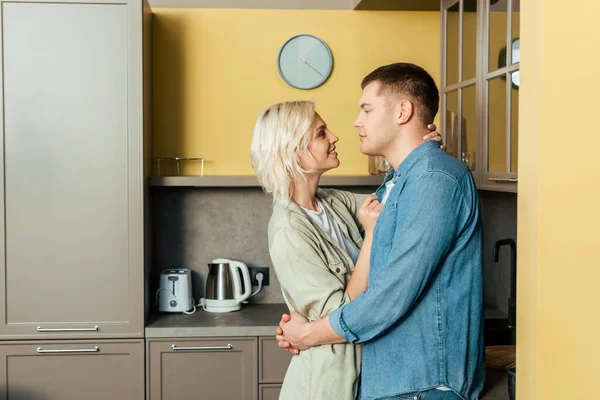 Vista lateral de sorrir jovem casal amoroso abraçando na cozinha em casa — Fotografia de Stock