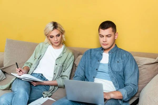 Joven pareja independiente en el sofá utilizando el ordenador portátil y la escritura en el cuaderno cerca de la pared amarilla - foto de stock