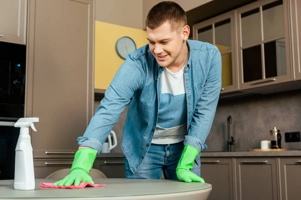 Улыбающийся мужчина в резиновых перчатках, моющий стол с тряпкой и моющим средством на кухне дома — стоковое фото