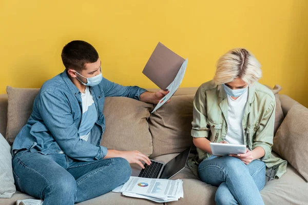 Jovem casal em máscaras médicas no sofá usando laptop e tablet digital perto de papéis durante a quarentena — Fotografia de Stock