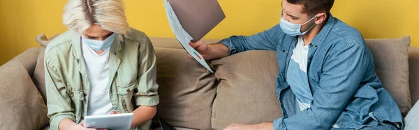 Jeune couple en masques médicaux sur canapé en utilisant une tablette numérique et des papiers de lecture pendant la quarantaine, orientation panoramique — Photo de stock