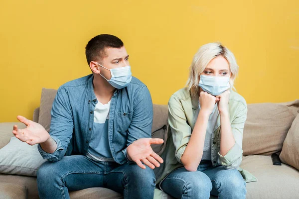 Jeune couple en masques médicaux se disputant sur canapé près du mur jaune — Photo de stock