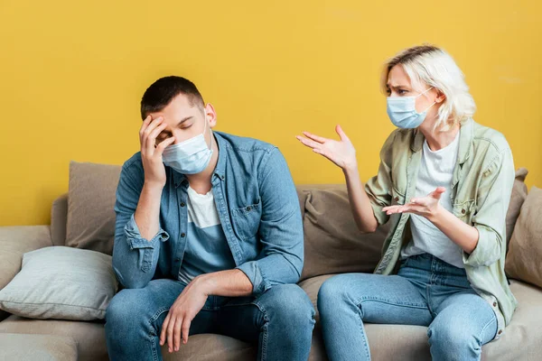 Young couple in medical masks quarreling on sofa near yellow wall — Stock Photo