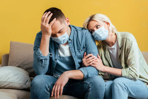 Joven pareja triste en máscaras médicas en sofá cerca de la pared amarilla - foto de stock