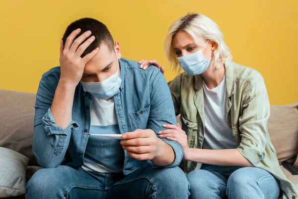 Triste giovane coppia in maschere mediche con termometro sul divano vicino alla parete gialla — Foto stock