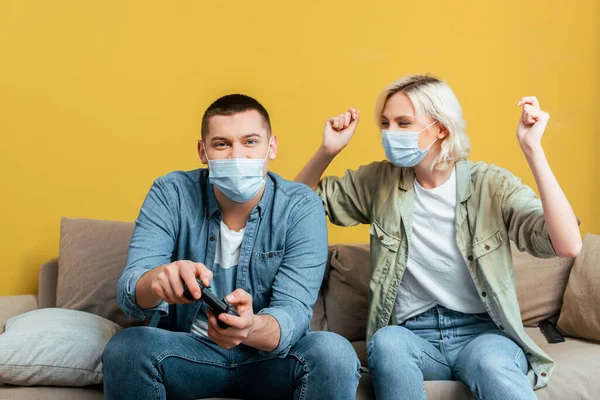 KYIV, UKRAINE - APRIL 22, 2020: young man in medical mask playing video games with cheerful girlfriend on sofa near yellow wall — Stock Photo