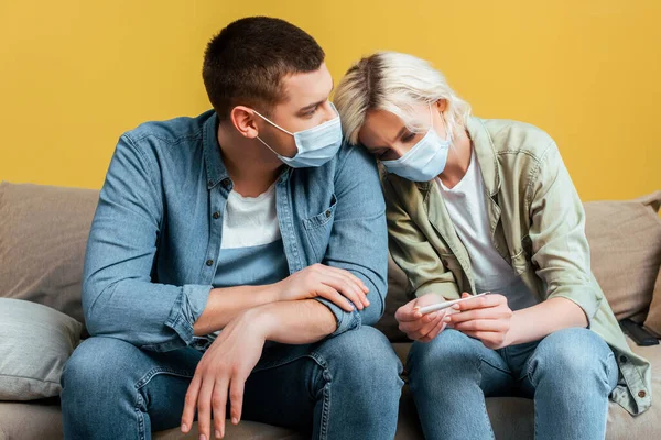 Triste pareja joven en máscaras médicas con termómetro en el sofá cerca de la pared amarilla - foto de stock