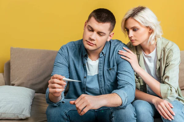 Sad young couple with thermometer on sofa near yellow wall — Stock Photo