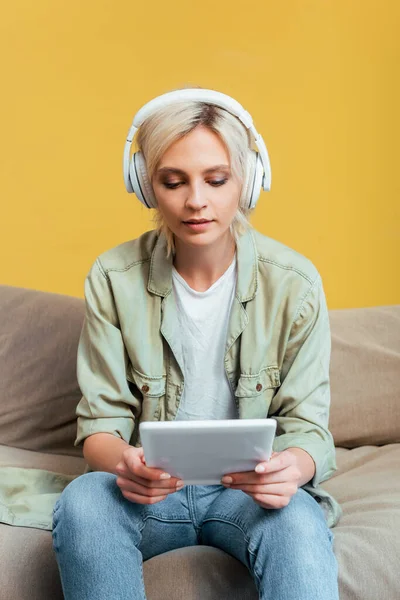 Young blonde woman in headphones with digital tablet on sofa near yellow wall — Stock Photo