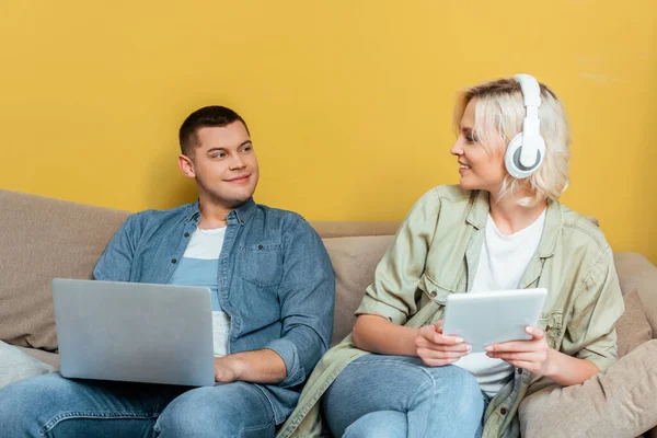 Smiling young blonde woman in headphones with digital tablet near boyfriend with laptop on sofa near yellow wall — Stock Photo