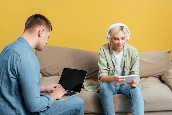 Smiling young blonde woman in headphones with digital tablet near boyfriend with laptop on sofa near yellow wall — Stock Photo