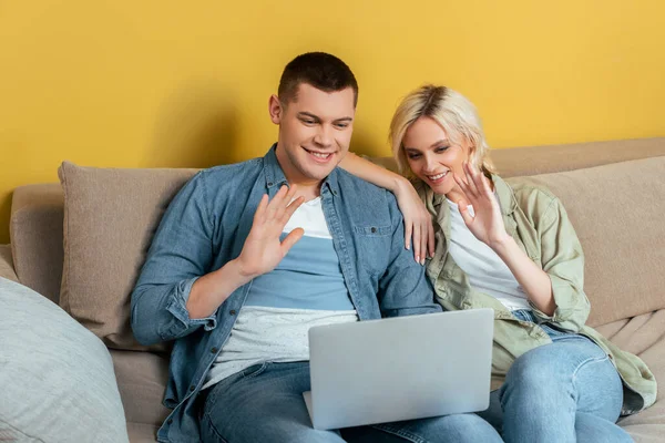 Feliz joven pareja en el sofá de vídeo chat en el ordenador portátil y saludando de la mano cerca de la pared amarilla - foto de stock