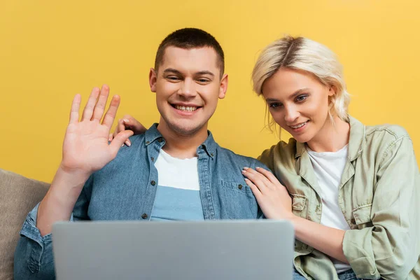 Счастливая молодая пара на диване с видео чатом на ноутбуке и размахивая рукой возле желтой стены — стоковое фото