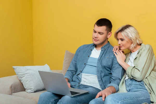 Молода пара на дивані сидить з ноутбуком біля жовтої стіни — стокове фото
