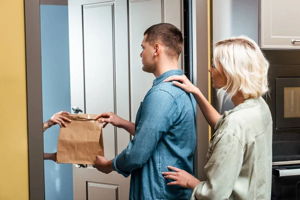 Delivery man giving package to young couple near open door at home — Stock Photo