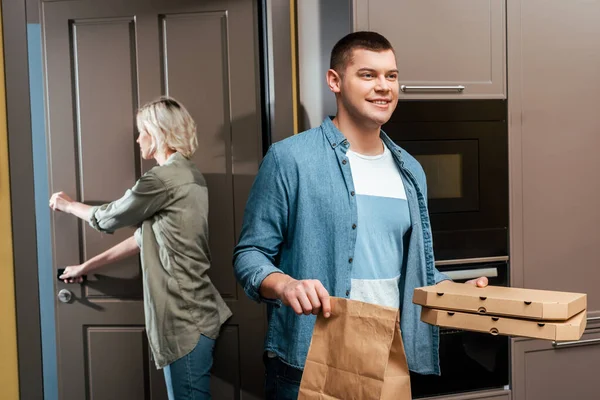 Улыбающийся мужчина с коробками для пиццы и пакетом рядом с девушкой закрывая дверь дома — стоковое фото