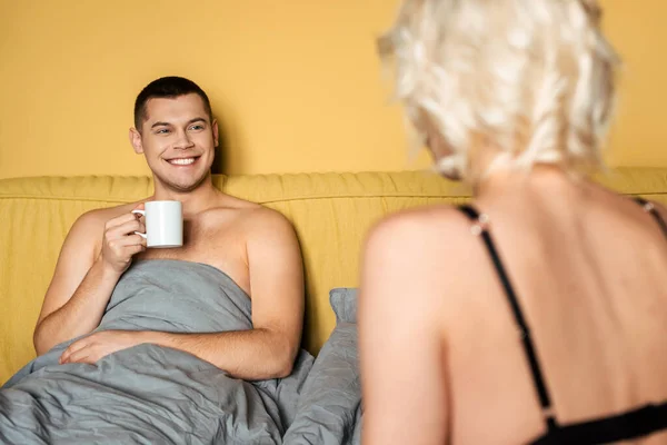 Selektiver Fokus eines lächelnden Mannes mit Becher, der neben blonder Frau im Bett liegt — Stockfoto