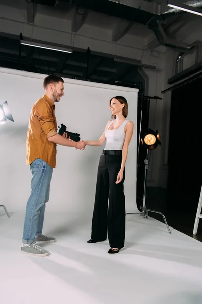 Vista lateral de la modelo sonriente estrechando la mano con el fotógrafo en el estudio de fotos - foto de stock