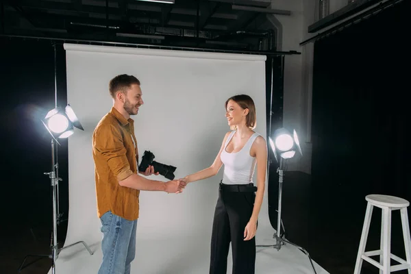 Vue latérale du photographe tenant un appareil photo numérique tout en serrant la main avec un modèle souriant en studio photo — Photo de stock