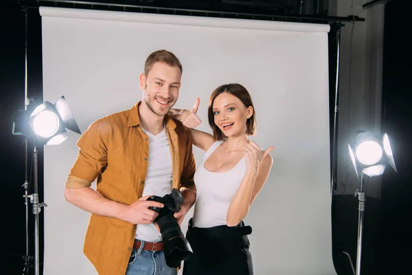 Modelo atraente mostrando polegares perto de fotógrafo sorridente com câmera digital no estúdio de fotos — Fotografia de Stock