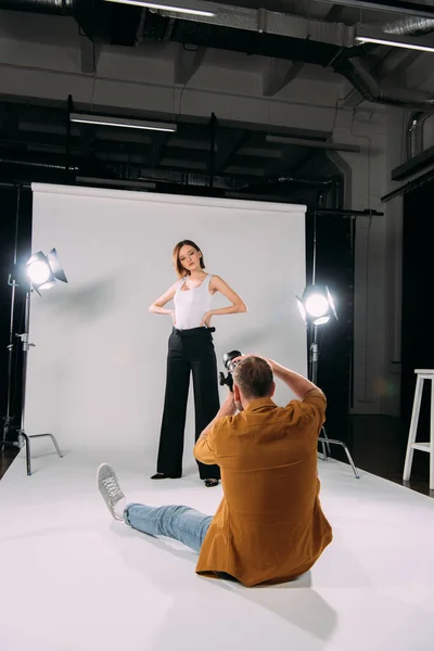Bella modella con le mani sui fianchi posa al fotografo seduto sul pavimento in studio fotografico — Foto stock