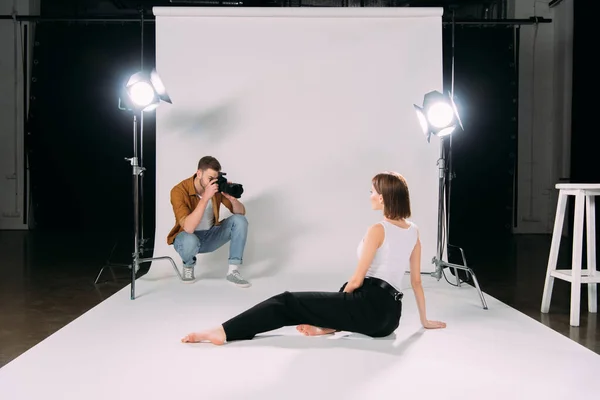 Fotógrafo sosteniendo la cámara digital mientras toma la foto de la joven modelo en el suelo en el estudio de fotos - foto de stock