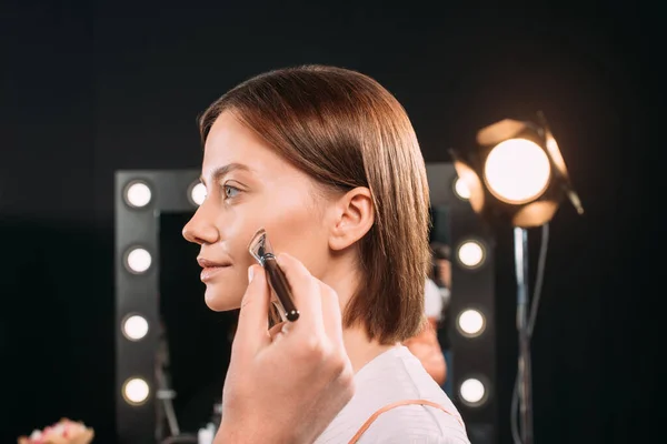 Maquillaje artista celebración de cepillo cosmético cerca de la cara de hermosa modelo en el estudio de fotos - foto de stock