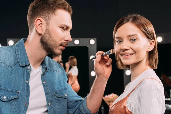 Beautiful model smiling at camera near makeup artist applying face foundation in photo studio — Stock Photo
