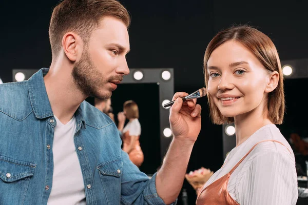 Hermosa modelo sonriendo a la cámara cerca de maquillaje artista aplicando la base de la cara en el estudio de fotos - foto de stock