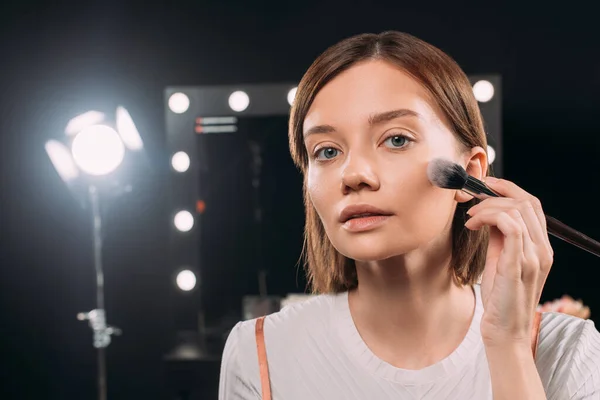 Donna attraente che tiene pennello cosmetico mentre applica la polvere per il viso in studio fotografico — Foto stock