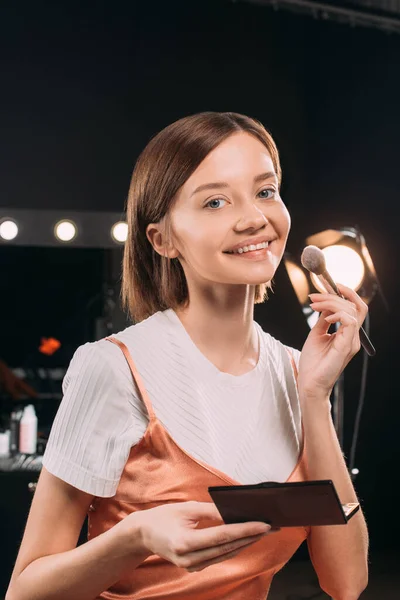 Beautiful woman smiling at camera while holding cosmetic brush and set of blush in photo studio — Stock Photo