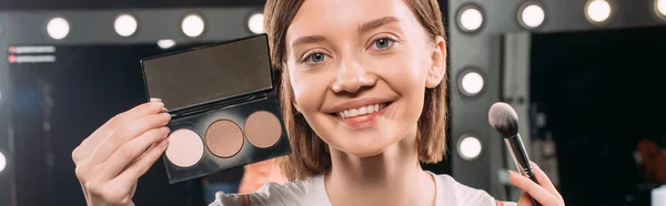 Recorte panorámico de niña sonriendo a la cámara mientras sostiene el conjunto de maquillaje y el cepillo cosmético en el estudio de fotos - foto de stock