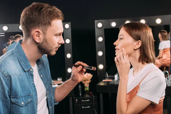 Vue latérale du modèle pointant sur la joue près du maquilleur tenant un pinceau cosmétique dans un studio photo — Photo de stock